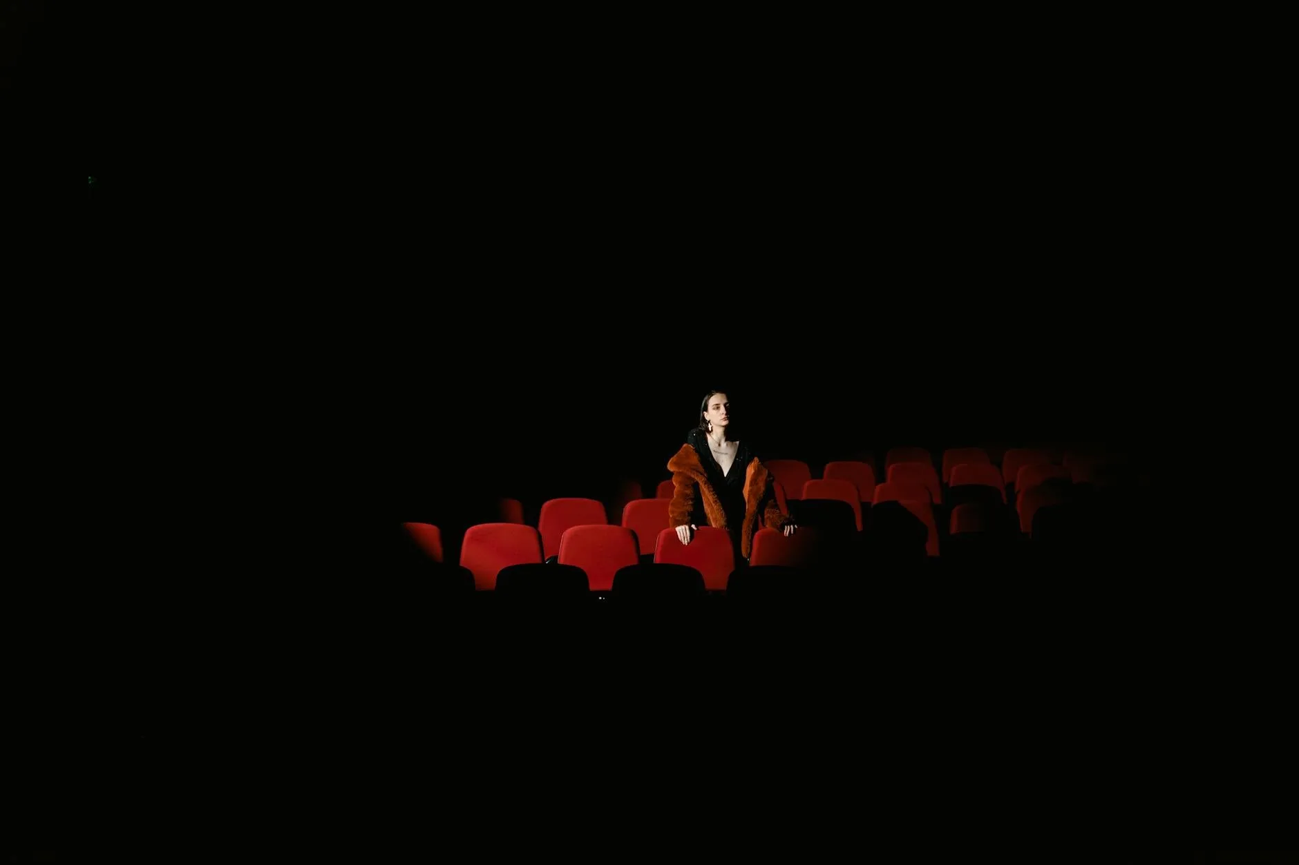 Kobieta w futrze siedzi samotnie w pustej, przyciemnionej sali teatralnej